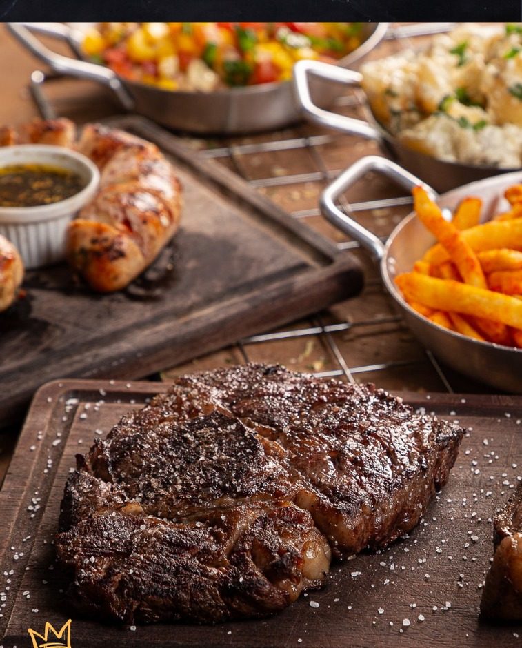 Steak ancho grelhado e salgado grosso sobre tábua de madeira, com fritas e farofa ao fundo.